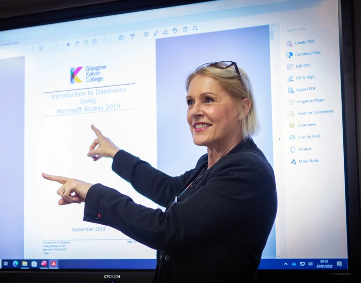 Lecturer in a black blazer presenting a lesson on Microsoft Access, pointing at a large interactive screen in a computer lab. Lecturer in a black blazer presenting a lesson on Microsoft Access, pointing at a large interactive screen in a computer lab.